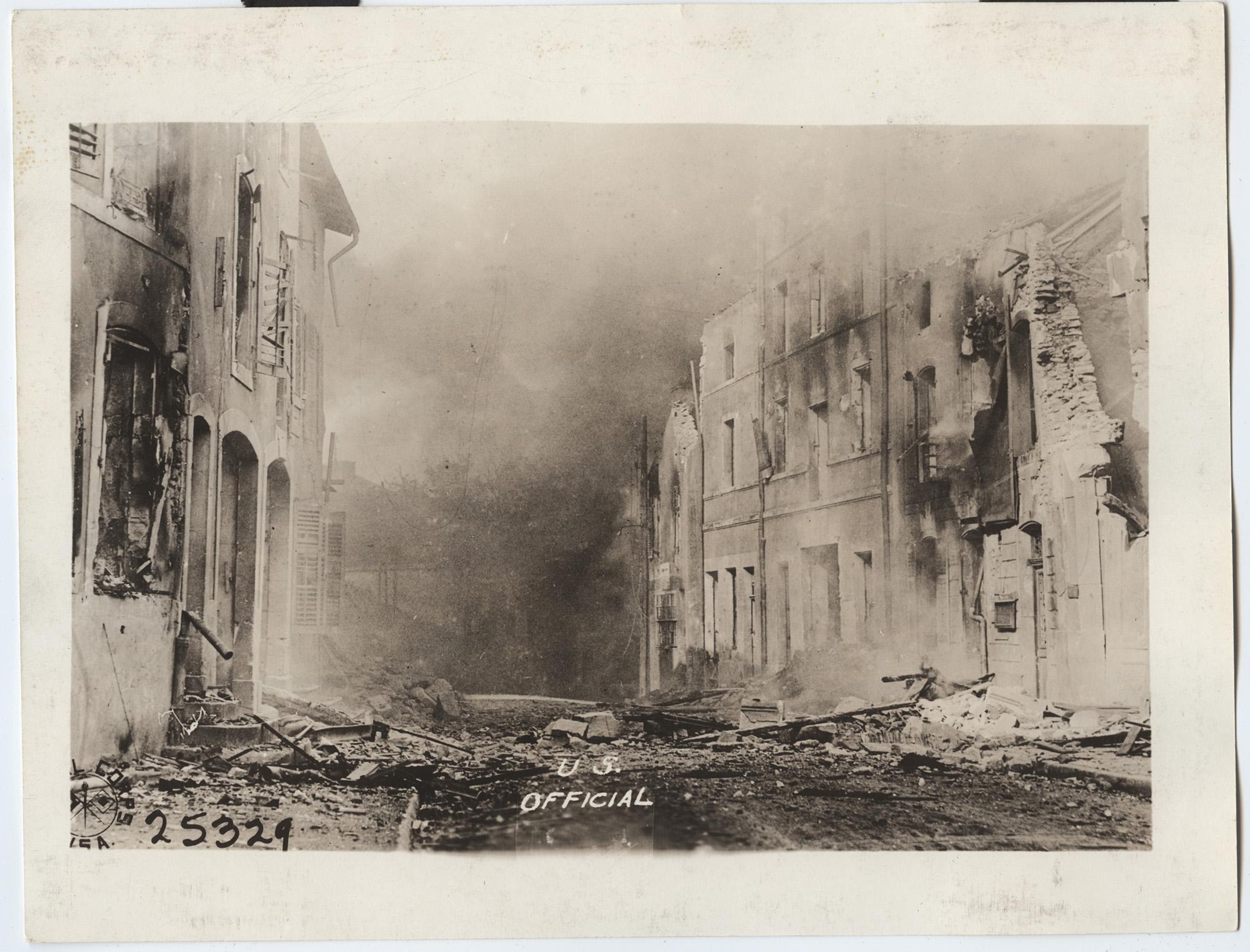 Black and white photograph of a street scene with destroyed buildings on either side and smoke in the air. Along the bottom of the image there text that says U.S. / OFFICIAL, another that says 25329, and a USA SIGNAL CORPS insignia.
