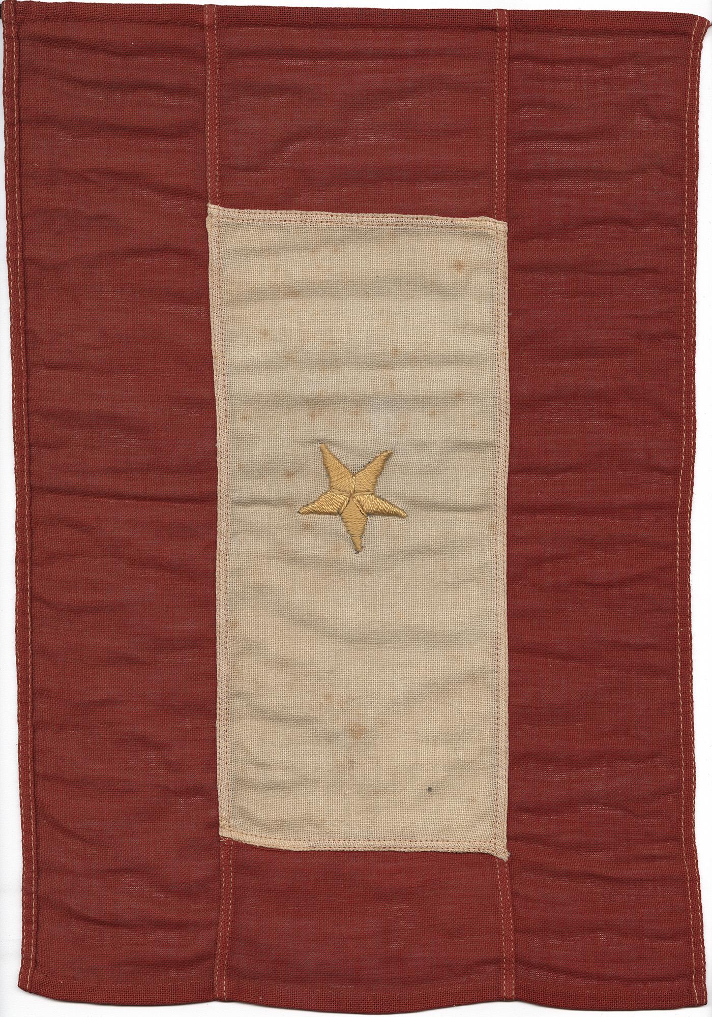 Cloth flag made of red fabric around the outside, white fabric in the middle, and a golden colored star embroidered into the center.