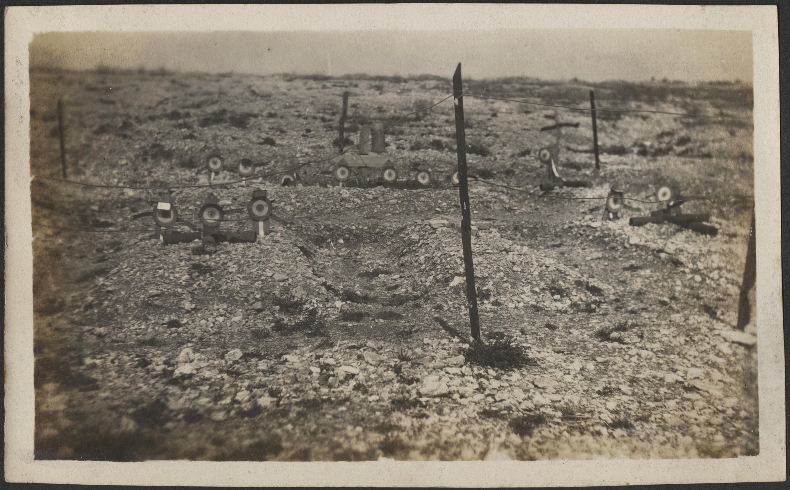 Black and white photograph of heavily pebbled ground with sticks stuck into the ground and rope between them, making a fence. Inside the fenced off area are ribbons stuck into the ground so that they stand up, acting as grave markers.