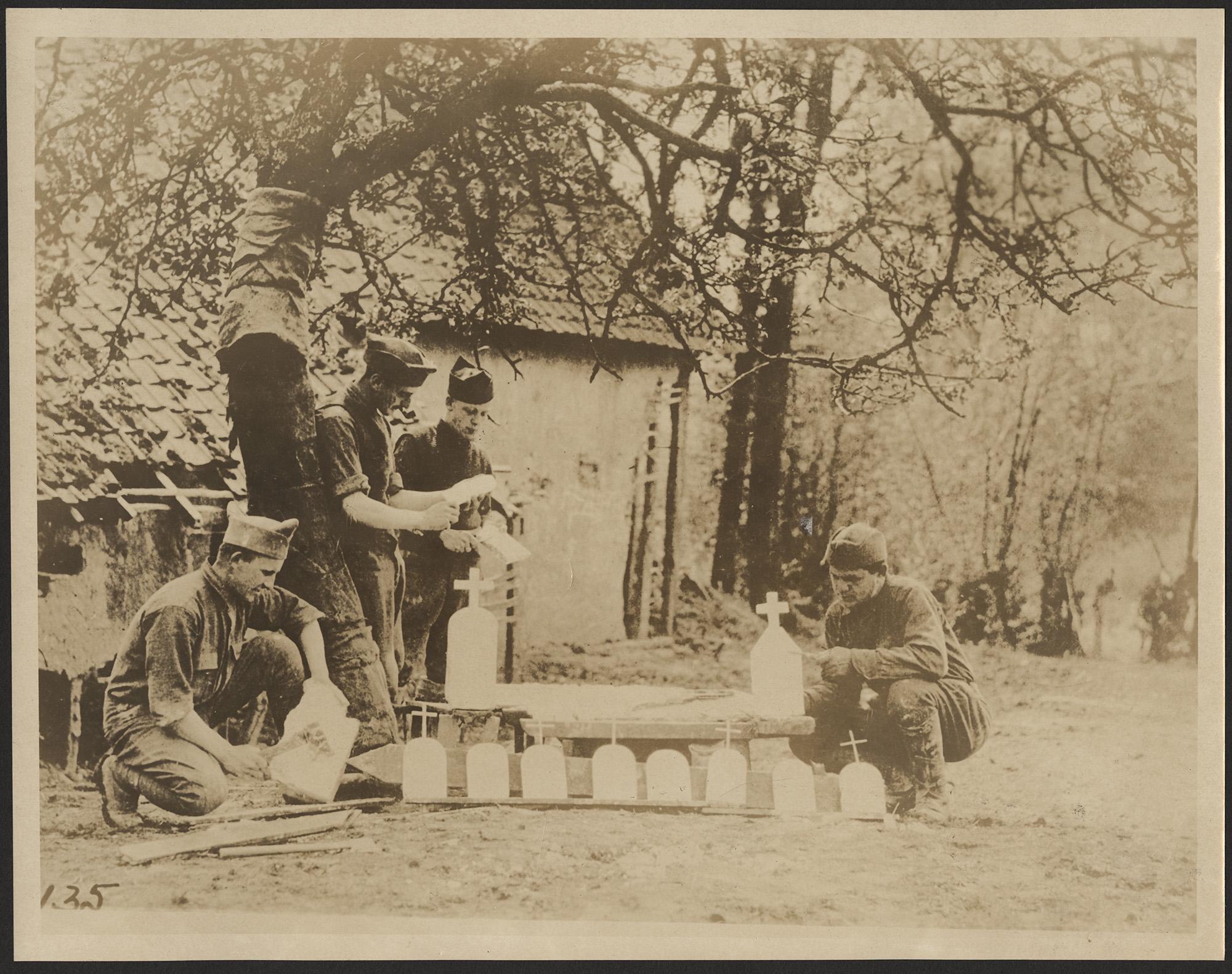 Black and white photograph with four men in uniform. Two are kneeling, one on either side of ten different small grave markers with crosses on top of them. Two men are standing in the center. Behind them is a building and trees. Each of the men has tools and unfinished markers in their hands.