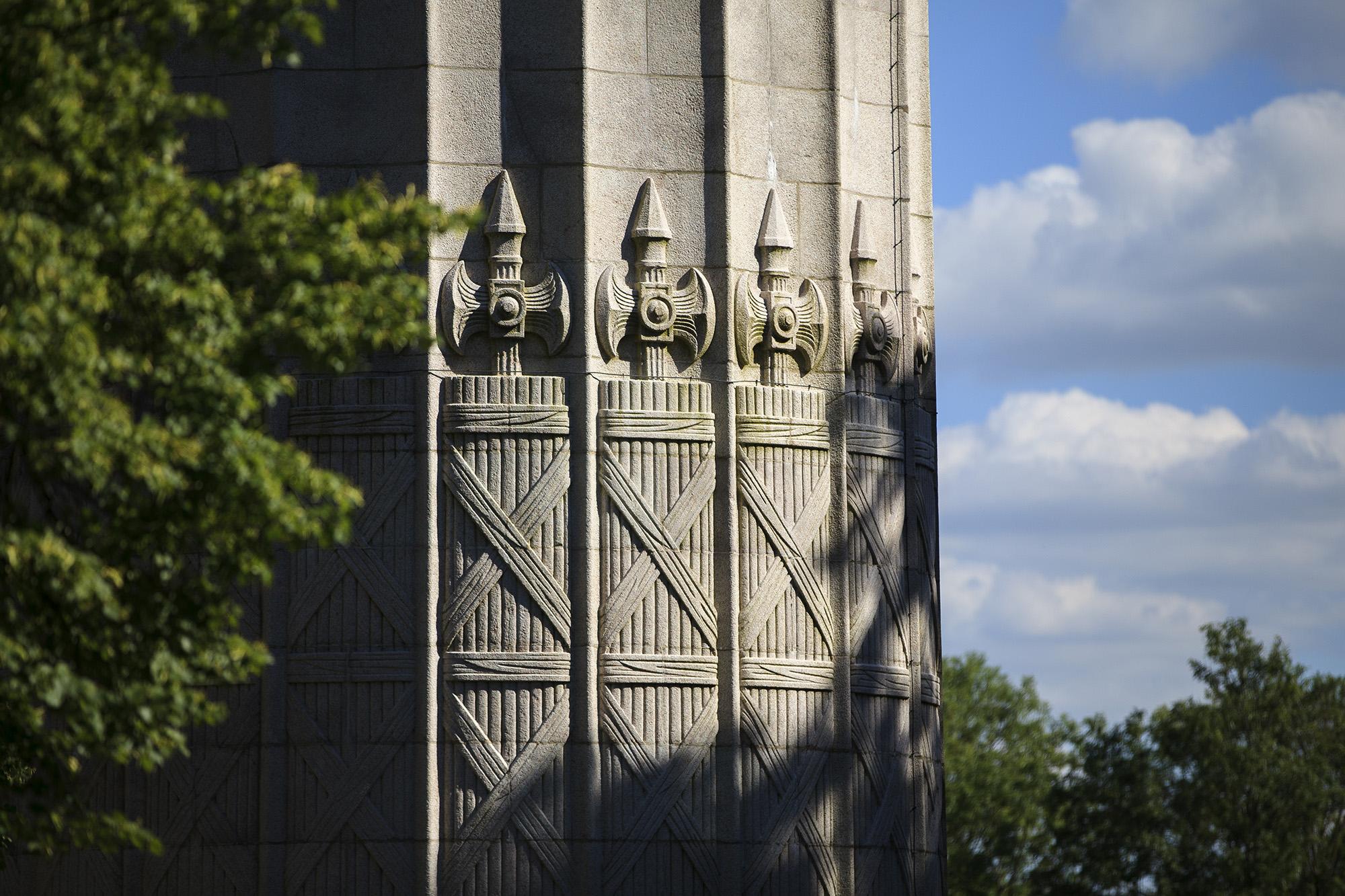 Modern color photo of the side of a massive stone column. The sides are carved with intricate depictions of battle axes.
