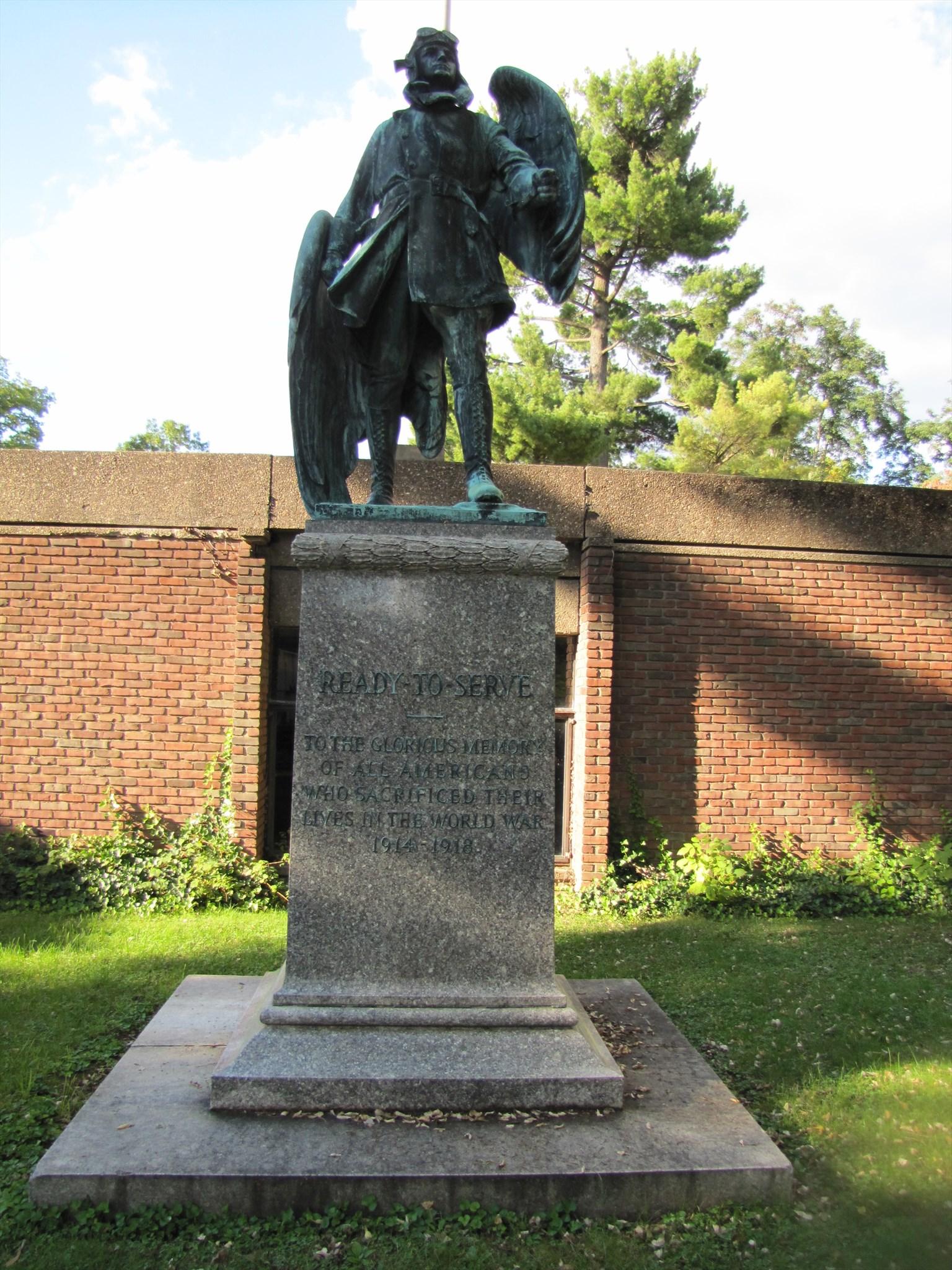 Color photograph of a statue of man in a pilot’s outfit with hat and goggles, with wings on his back. The statue sits atop a pedestal with an inscription. Behind the statue is a brick building and the statue sits in a grassy area.