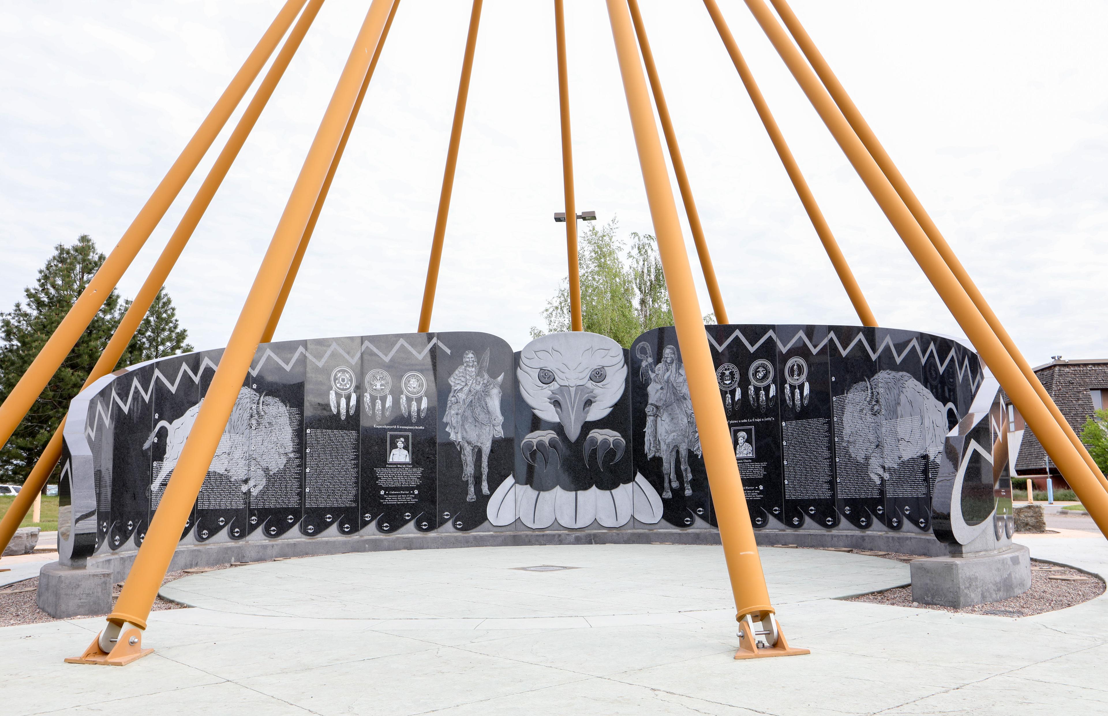 Color photograph of a curved wall engraved with text and images of bison, people on horseback, individual soldiers, and other imagery. The center of the wall features the body of an eagle with its wings acting as the rest of the wall. There are yellow-orange poles around it on the ground, going up and meeting above the wall. The ground is paved and there are trees and a building in the background.
