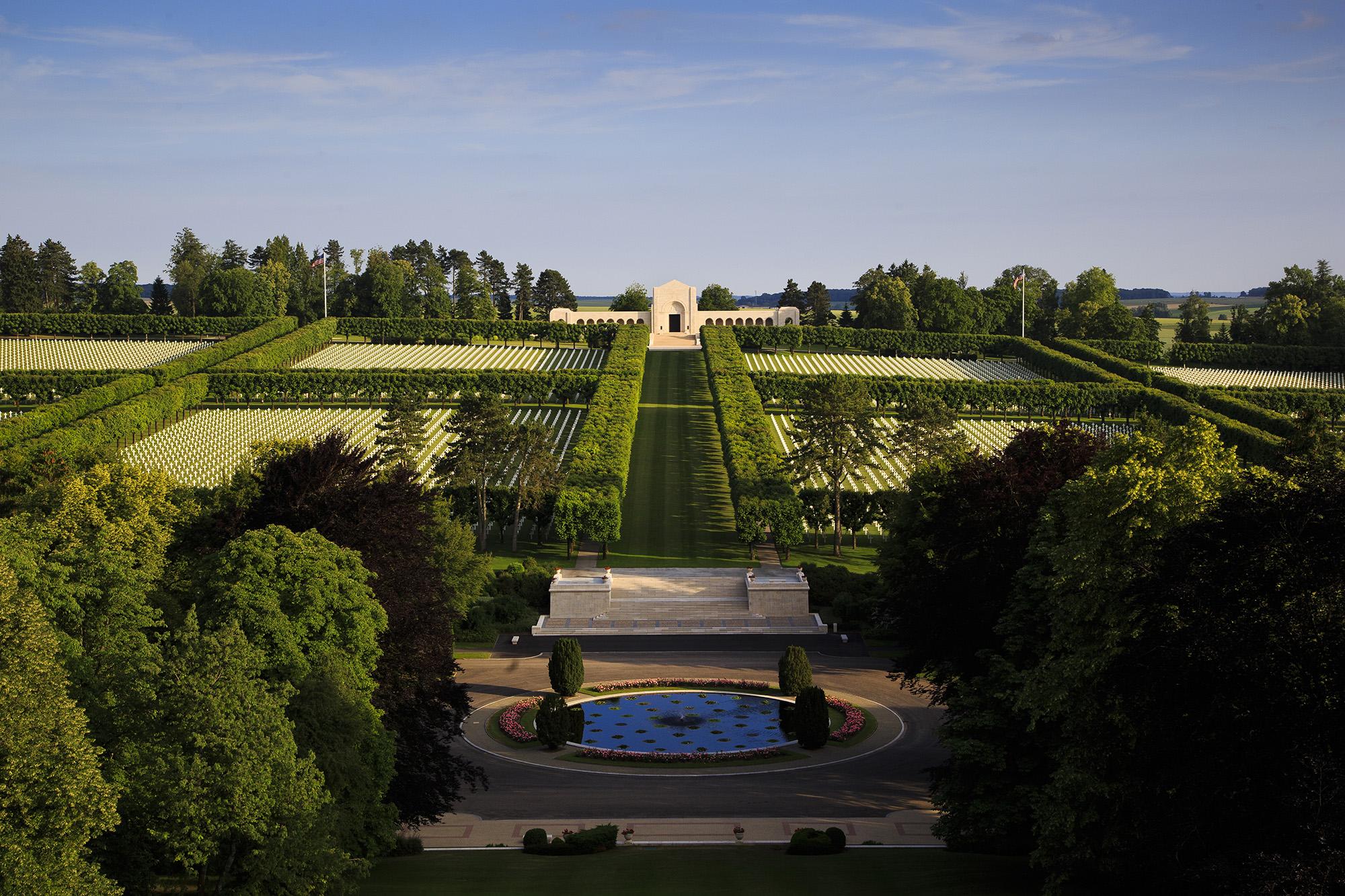 Color photograph of a large cemetery with white headstones and large hedges sectioning off a variety of areas. In the foreground are trees and a pond with flowers and bushes. In the background, there is a white building with archways, a large door and stairs leading up to it, along with trees and two American flags on poles.