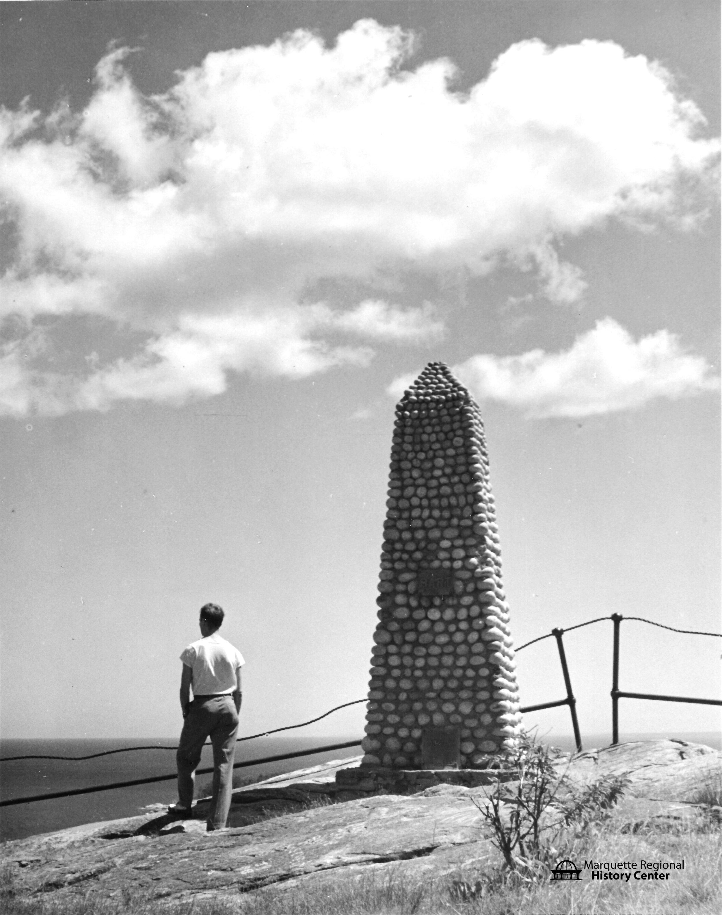 Black and white photograph on the top of a cliffside with light clouds in the sky. In the front center a man in a white shirt and darker pants stands, looking away from the camera and to the water and sky in the background. To his right, there is an obelisk about twice his height made of stones. There are a few plants on the ground and there is a fence on the edge of the cliff.