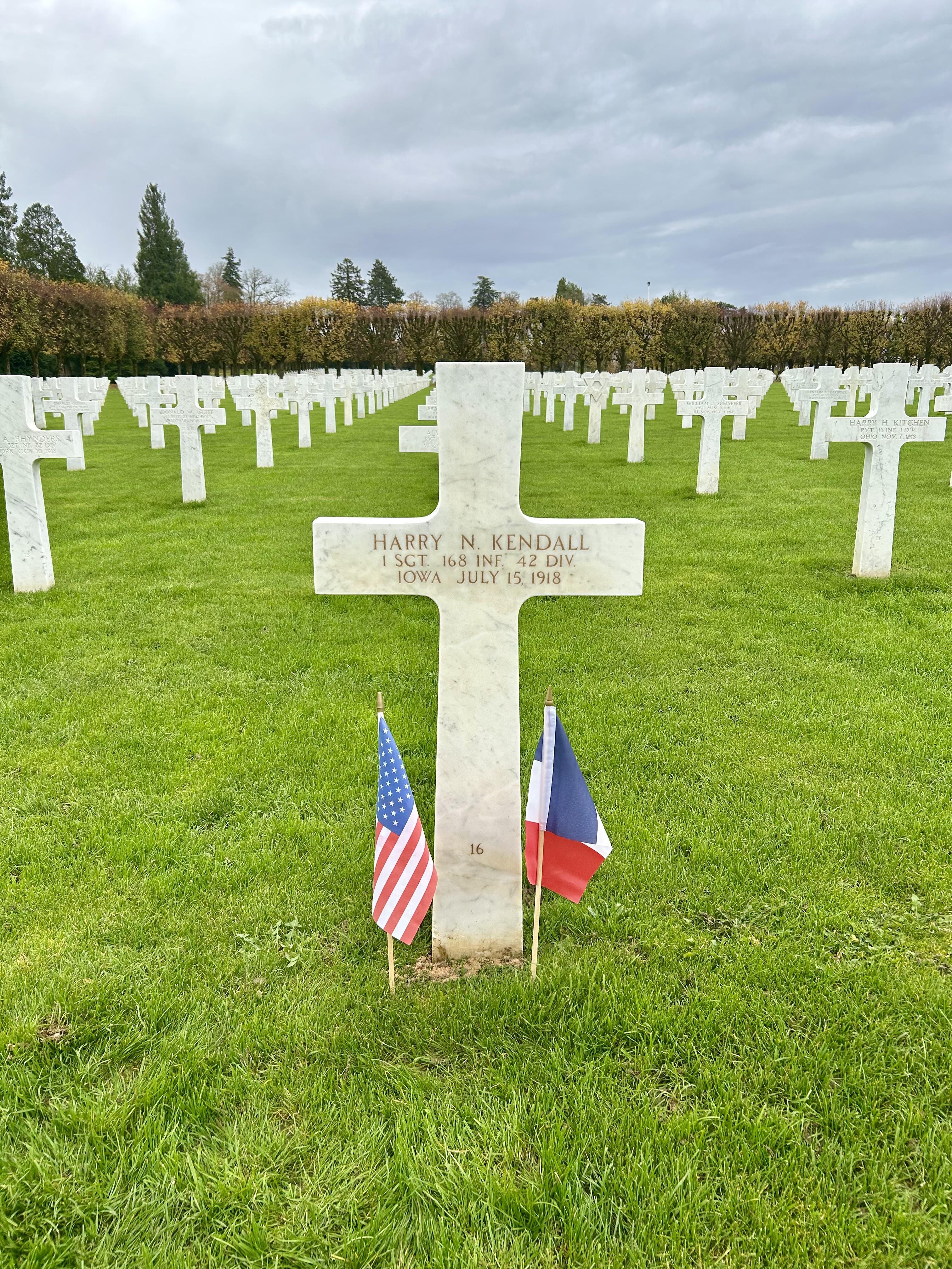 A color photograph of a grassy landscape with rows of white crosses in the ground. In the background stand many trees and there is a white cross in the ground at the center of the image, and small American and French flags in the ground on either side of the cross. The grave reads HARRY N. KENDALL / 1 SCT. 168 INF. 42 DIV. / IOWA JULY 15, 1918.
