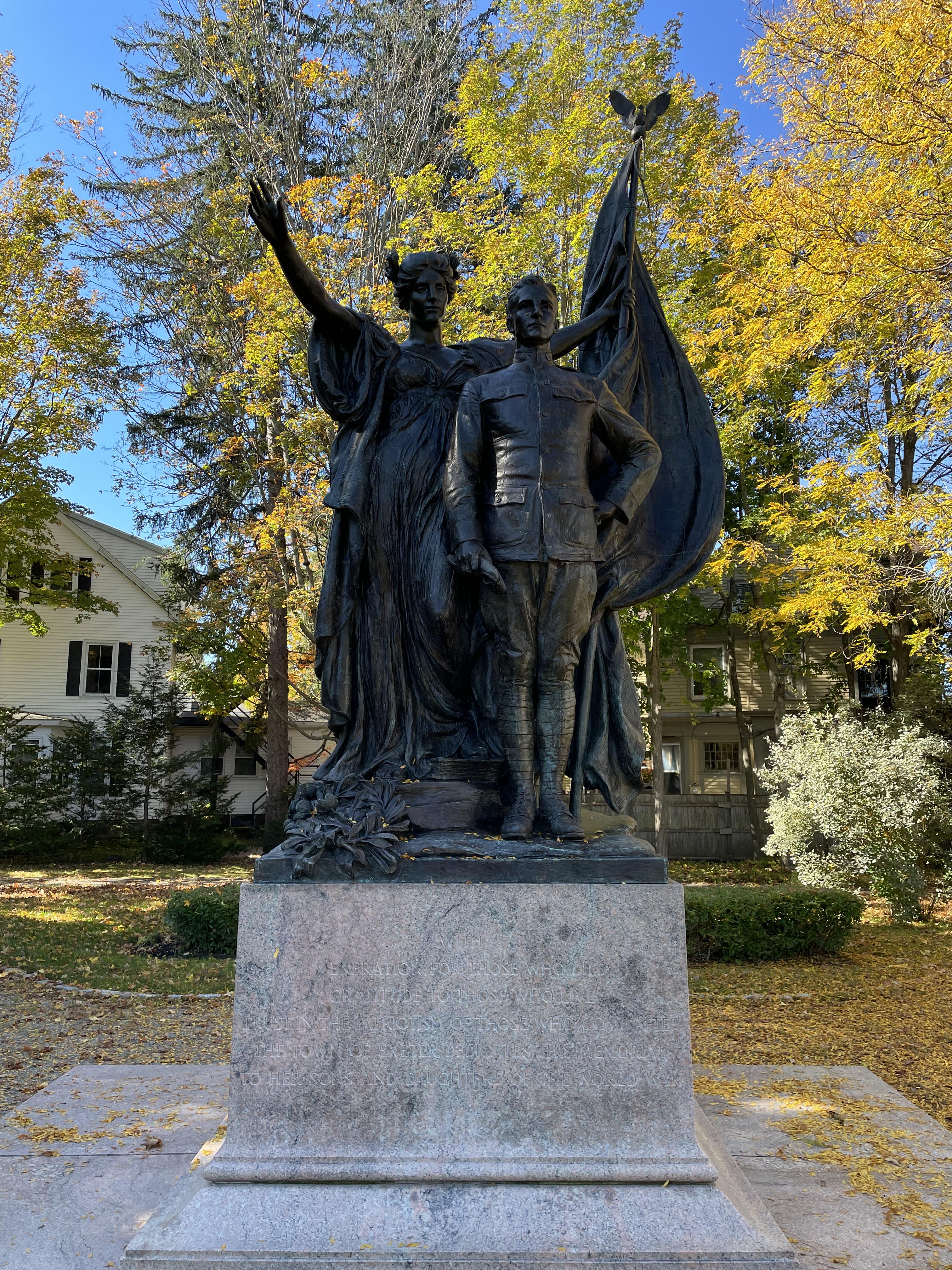 Color photograph of a statue of a woman with her arms outstretched, in a long flowing dress, with an American flag in one hand, which is behind a soldier standing with his hat in his hand. The statue sits atop a pedestal in a grassy area. In the background are trees, bushes and two buildings.