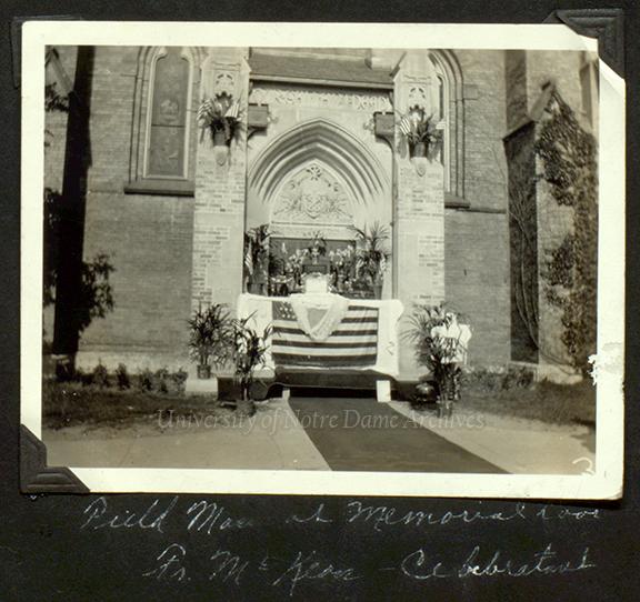 Black and white photograph of an ornately decorated doorway into a building. In front of the stone building, there is a table with an American flag and many flowers and plants in vases.