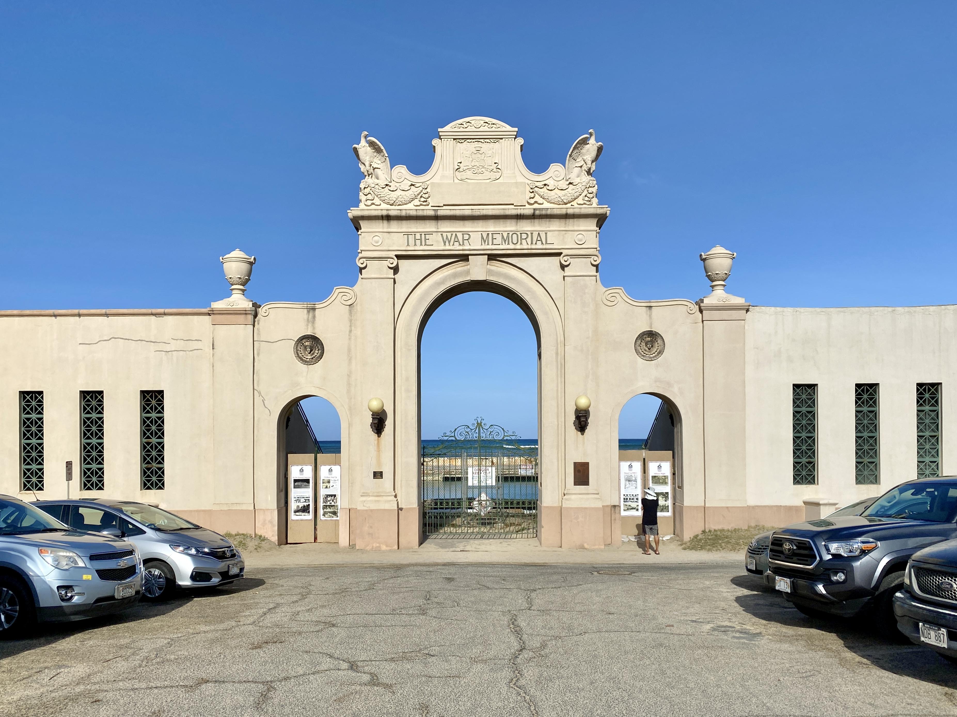 Color photo of a gate made of stone with an ornate top portion, including eagles and symbols. At the center bottom, there is a closed metal gate with water that can be seen through it on the other side. A person stands to the right of the gate, reading posters on the wall. There are cars in the foreground and a blue sky.